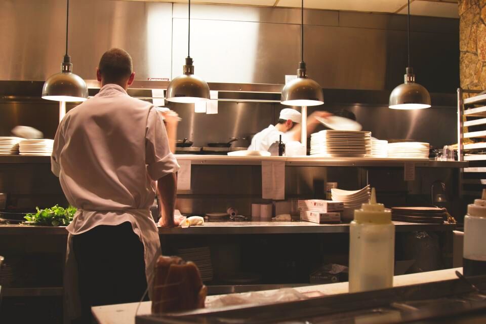 Cozinha profissional vista de fora, com bancadas inox e cozinheiros ao fundo